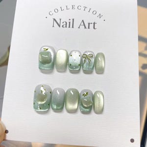 May include: A set of ten green and white ombre nail tips with various designs, including a bow, a star, and a leaf. The nail tips are displayed on a white card with the text "COLLECTION. Nail Art."