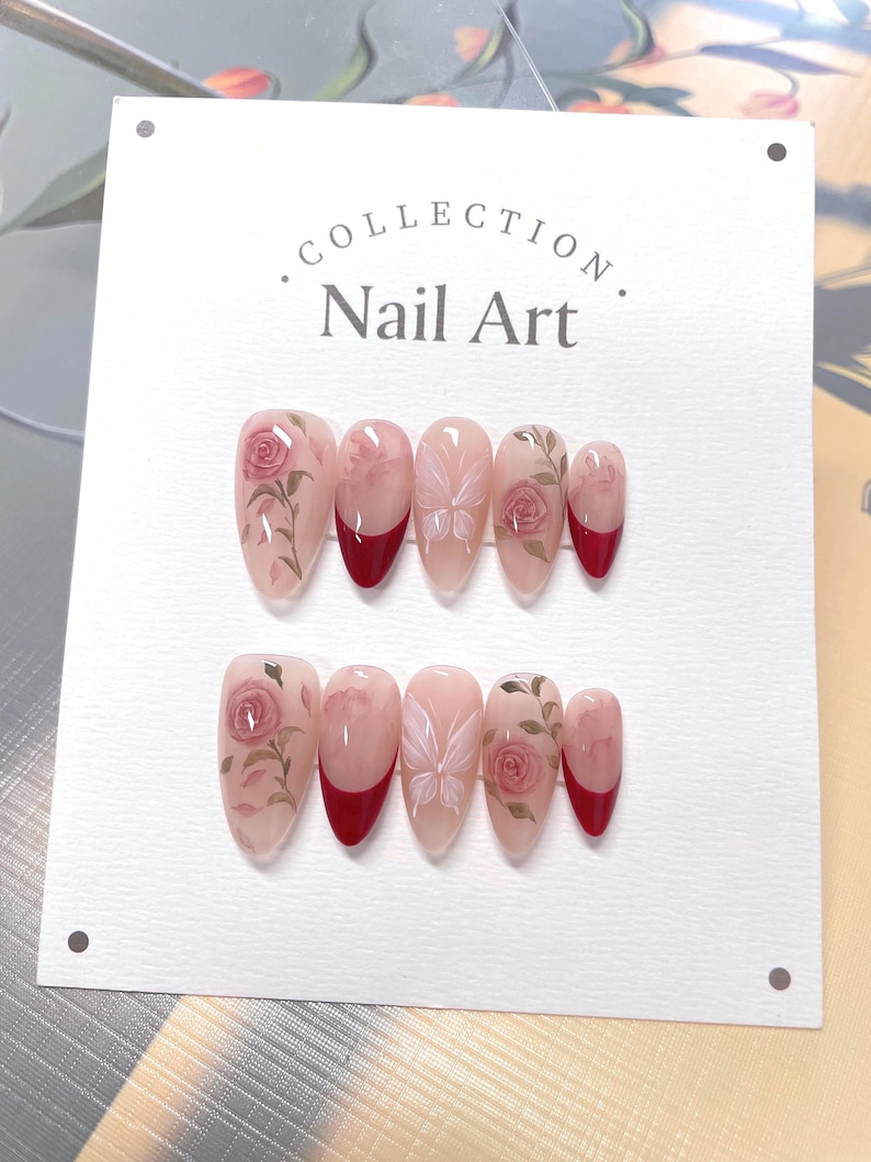 May include: A set of ten fake nails with a floral design. The nails are a light pink color with a red tip. The floral design features pink roses, green leaves, and white butterflies.