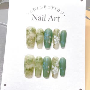 May include: A set of ten green and white nail art tips with a floral design. The tips are arranged in two rows of five. The tips are on a white background with the text "COLLECTION. Nail Art." printed on the top.