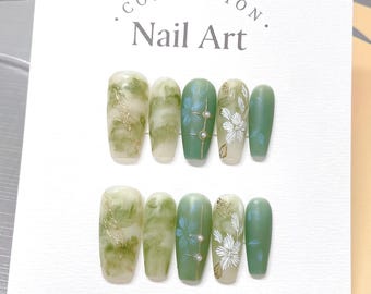 May include: A set of ten green and white nail art tips with a floral design. The tips are arranged in two rows of five. The tips are on a white background with the text "COLLECTION. Nail Art." printed on the top.