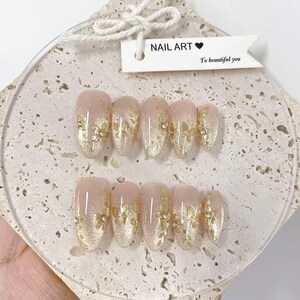 May include: Set of oval-shaped false nails with a nude base and gold floral designs. Each nail is adorned with gold glitter and small rhinestones. The tag reads "NAIL ART" and "To beautiful you".