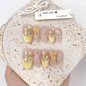 May include: A set of artificial nails with a floral design. Each nail has a light pink base with a yellow tip and a painted floral arrangement in pink, purple, and green. Some nails feature a yellow bow design. The tag reads "NAIL ART" and "To beautiful you."