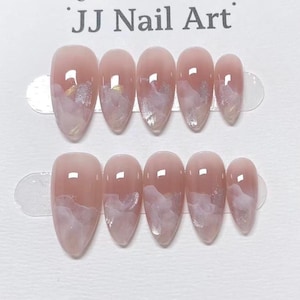 May include: Two sets of almond-shaped false nails. Each nail is a light pink with a white and gold cloud design. The text "JJ Nail Art" is at the top.