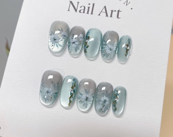 May include: A set of six light blue oval-shaped artificial nails with white floral designs and gold accents. The nails are displayed on a white card with the text "COLLECTION. Nail Art."