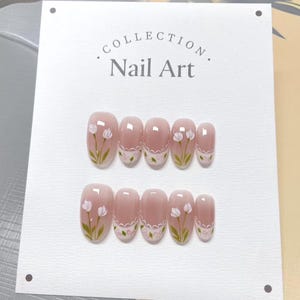 May include: A set of ten nude-colored oval-shaped fake nails with white flowers and green leaves painted on them. The nails are arranged in two rows on a white card with the text "COLLECTION. Nail Art." printed on it.