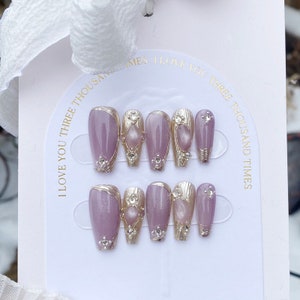 Peut inclure: Ensemble de dix faux ongles avec une base violet clair et des accents argent&eacute;s. Chaque ongle a un motif argent&eacute; avec un strass transparent au centre. Les ongles sont dispos&eacute;s en deux rang&eacute;es sur un fond blanc. Le texte "I love you three thousand times" est imprim&eacute; sur le fond.