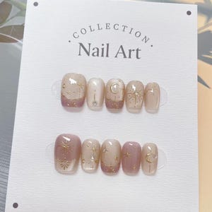 May include: A set of false nails with a celestial theme. The nails are in light pink and nude shades, decorated with gold star and moon designs. The card displays the text "COLLECTION Nail Art".