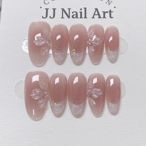 Può includere: Due set di unghie finte con una sfumatura rosa chiaro. Ogni unghia presenta un motivo floreale bianco e dettagli dorati. Il testo "JJ Nail Art" è visibile in alto. Le unghie sono di forma ovale e sembrano pronte per l'applicazione.