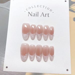Può includere: Un set di unghie finte con un design a marmo rosa chiaro e bianco. Ogni unghia ha un top coat trasparente e una piccola gemma decorativa. Le unghie sono esposte su una carta bianca con la scritta "COLLECTION Nail Art".