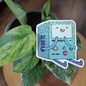 May include: A holographic sticker of a green and blue cartoon robot with a happy face. The robot has a screen, buttons, and a joystick. The sticker is on a green plant.