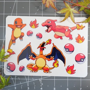 May include: A white rectangular sticker sheet featuring cartoon images of Charmander, Charmeleon, and Charizard, along with red and white Poké Balls and flames. The characters are orange, red, and yellow with black outlines. The sticker sheet is on a gray surface.