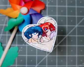 May include: A heart-shaped sticker with a cartoon illustration of two women with blue and red hair, sleeping together in a bed. The sticker has a sparkly, iridescent finish.