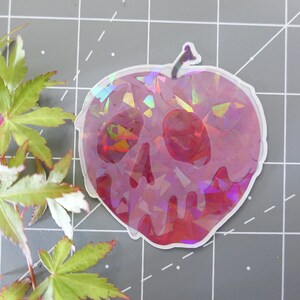 May include: A holographic sticker of a red apple with a skull design. The apple is dripping with a red liquid. The sticker is on a gray background.