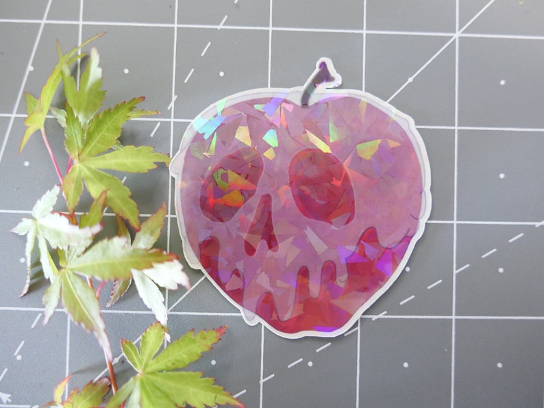 May include: A holographic sticker of a red apple with a skull design. The apple is dripping with a red liquid. The sticker is on a gray background.