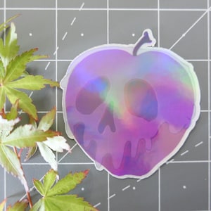 May include: A holographic sticker of a purple apple with a skull face. The apple is dripping with purple liquid.