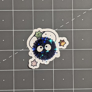 May include: A black and blue iridescent sticker of a round, fuzzy creature with large eyes and a star on top. The creature is surrounded by smaller stars.