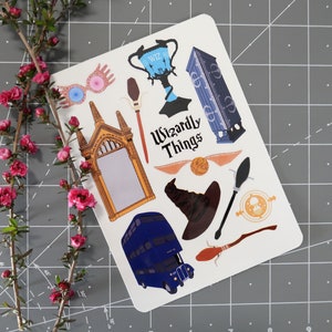 May include: A white sticker sheet with various Harry Potter themed illustrations, including a Hogwarts castle, a golden snitch, a broomstick, a sorting hat, and a double-decker bus. The text "Wizardly Things" is printed in black on the sticker sheet.