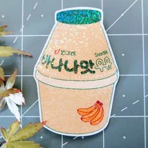 May include: A sticker of a banana milk carton with a glittery finish. The carton is light brown with a green lid and a green label that says "Banana Milk" in Korean. The label also says "Since 1974". There are three yellow bananas on the bottom of the carton.