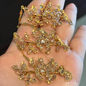May include: Three gold-toned hair clips with floral and leaf designs, adorned with numerous clear and pink gemstones. The clips are connected by delicate chains. The image includes the text "HALIM ANTIQUE JEWELRY".