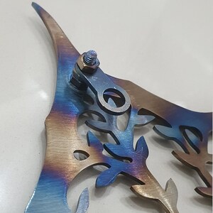 Puede incluir: Una pieza de arte de pared de metal en forma de &aacute;rbol con un acabado iridiscente azul, morado y dorado. El &aacute;rbol tiene un dise&ntilde;o detallado con muchas hojas y ramas. La pieza tiene un anillo de metal unido a la parte superior para colgar.