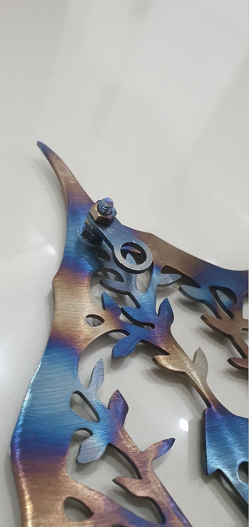 Puede incluir: Una pieza de arte de pared de metal en forma de &aacute;rbol con un acabado iridiscente azul, morado y dorado. El &aacute;rbol tiene un dise&ntilde;o detallado con muchas hojas y ramas. La pieza tiene un anillo de metal unido a la parte superior para colgar.