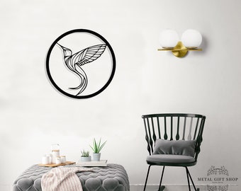 Peut inclure: Décoration murale en métal noir représentant un colibri dans un cadre circulaire. L'œuvre est exposée sur un mur blanc avec un pouf gris et un fauteuil à bascule noir au premier plan.