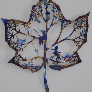 Puede incluir: Una escultura de pared de metal con forma de hoja de arce azul y marr&oacute;n con recortes de p&aacute;jaros, una casita de p&aacute;jaros, una ni&ntilde;a en bicicleta y un gato.
