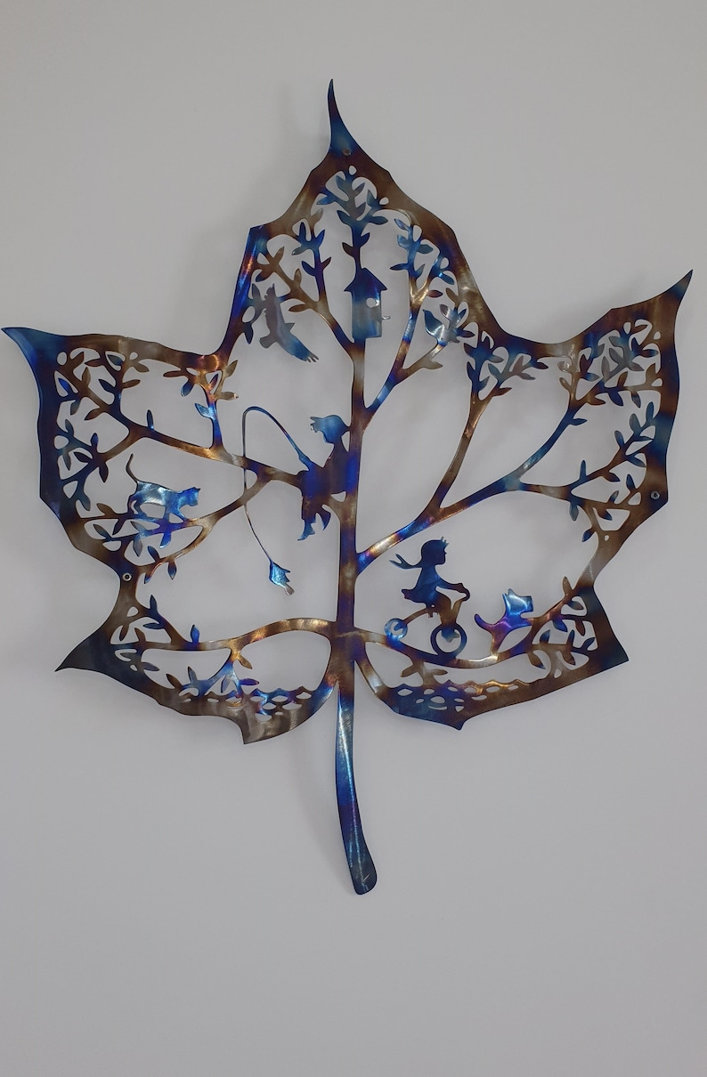 Puede incluir: Una escultura de pared de metal con forma de hoja de arce azul y marr&oacute;n con recortes de p&aacute;jaros, una casita de p&aacute;jaros, una ni&ntilde;a en bicicleta y un gato.