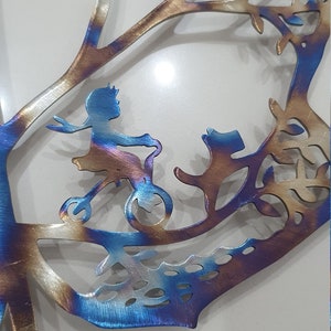 Puede incluir: Una pieza de arte de pared de metal que representa a una ni&ntilde;a montando un triciclo en un &aacute;rbol. El arte est&aacute; hecho de metal y tiene una combinaci&oacute;n de colores azul, dorado y marr&oacute;n.