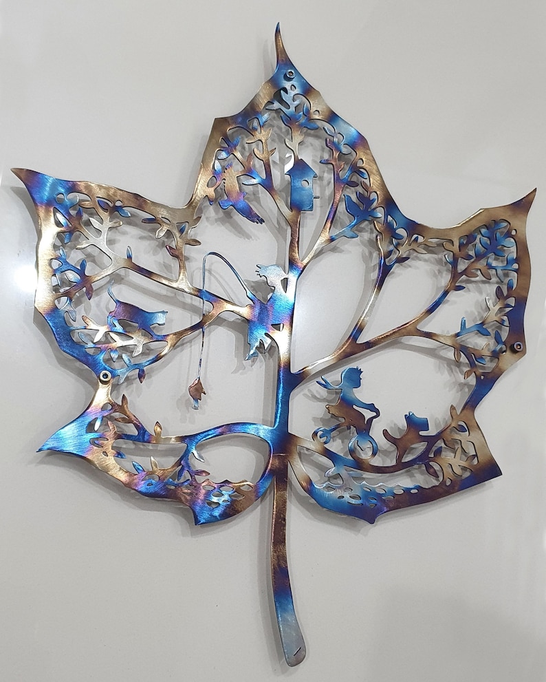 Puede incluir: Una escultura de pared de metal con forma de hoja de arce con un dise&ntilde;o detallado de un &aacute;rbol con p&aacute;jaros, una casita de p&aacute;jaros y un hada montando una bicicleta. La hoja est&aacute; pintada en tonos de azul, dorado y marr&oacute;n.