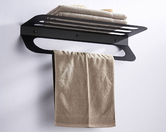 Puede incluir: Toallero de metal negro con un estante y una barra para toallas. El toallero está montado en una pared blanca y tiene una toalla colgando de la barra.