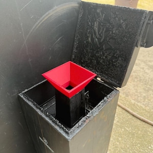 May include: A red plastic funnel attachment sits atop a black metal Masterbuilt gravity-fed charcoal smoker hopper. The funnel has a square opening and a tapered neck, positioned inside the open rectangular hopper lid of the smoker unit.
