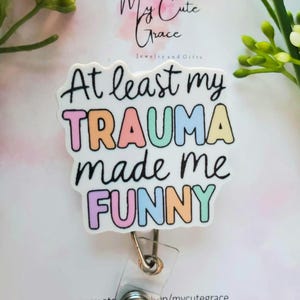 Peut inclure: Un badge blanc avec le texte "At least my TRAUMA made me FUNNY" en noir et couleurs pastel. Le badge a une sangle en plastique transparent et un clip en métal. Le fond comprend un motif floral et le texte "My Cute Grace Jewelry and Gifts".