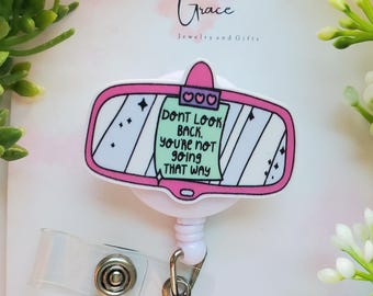 May include: A pink and white badge reel with a rearview mirror design. The mirror has the text "DON'T LOOK BACK, YOU'RE NOT GOING THAT WAY." The reel has a clear plastic strap and a metal clip. The Etsy shop URL is visible.