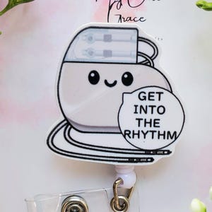 May include: A white and black badge reel featuring a cartoon pacemaker design with a smiling face. The badge reel has the text "GET INTO THE RHYTHM" in a speech bubble. The reel has a clear plastic strap and a metal clip.