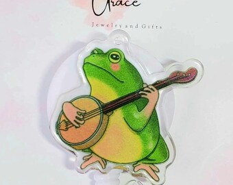May include: A green frog playing a banjo with a pink blush on its cheeks. The frog is on a white background with a clear plastic retractable badge holder.