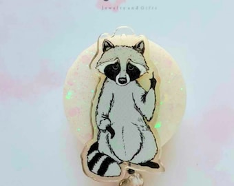 May include: A clear acrylic badge reel with a white glitter background. The reel features a cartoon raccoon with a black and white striped tail, sitting with its right paw raised in a middle finger gesture. The text "My Cute Grace Jewelry and Gifts" is printed on the background.
