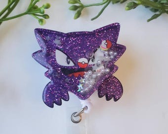 May include: Purple glitter resin badge holder featuring a cartoon image of the Pokemon, Gengar. The holder has a clear plastic backing and a retractable badge reel.