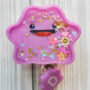 May include: A pink glitter resin badge holder featuring a cartoon character with a smiling face. The holder has a clear plastic badge holder attached to it with a purple glitter charm with the letter "D" on it.