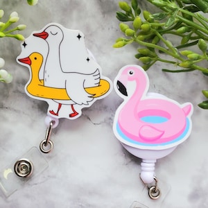 Puede incluir: Dos portatarjetas retráctiles con diseños de dibujos animados. Uno presenta un ganso blanco y un pato amarillo en una boya amarilla. El otro es un flamenco rosa en una boya azul y blanca. Ambos tienen una correa de plástico transparente y un clip de metal.
