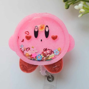 May include: Pink resin badge holder featuring a cartoon character with a pink face, black eyes, and red hearts. The holder has a clear resin section filled with glitter, hearts, stars, and flowers.