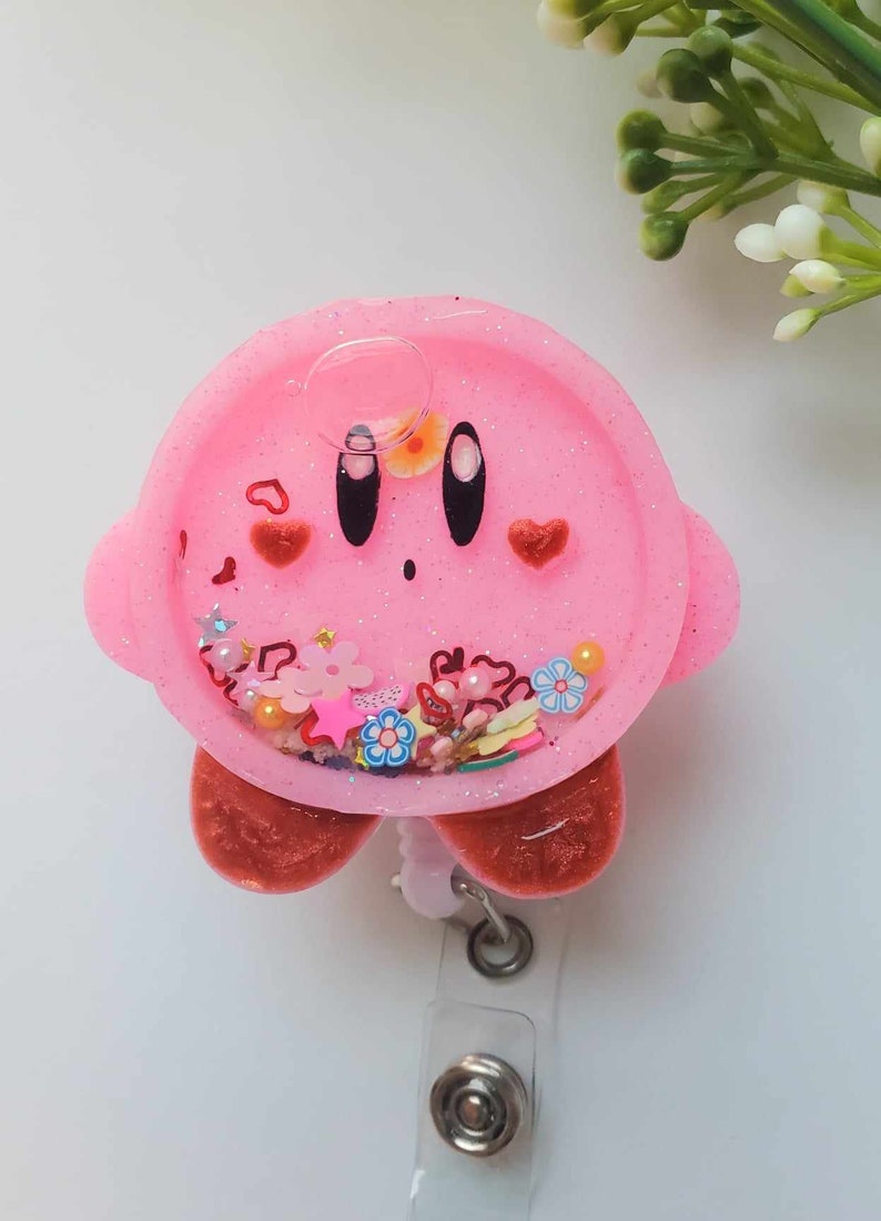 May include: Pink resin badge holder featuring a cartoon character with a pink face, black eyes, and red hearts. The holder has a clear resin section filled with glitter, hearts, stars, and flowers.