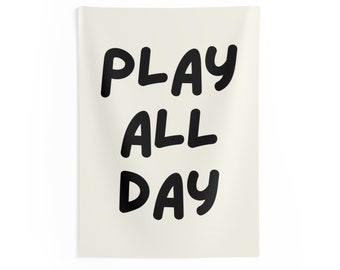 May include: A rectangular wall hanging with the words "PLAY ALL DAY" in bold, black, rounded letters. The background is a light cream colour. The wall hanging is suspended on a white wall.