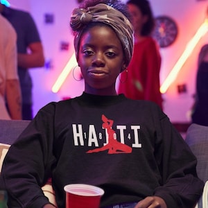 May include: Black sweatshirt with the word "HAITI" in white block letters and a red silhouette of a person. The sweatshirt has a high neckline and is worn by a person with a patterned head wrap and hoop earrings.