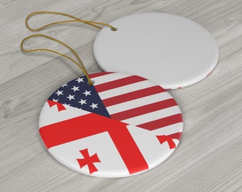 May include: Two white ceramic ornaments with gold string loops. One ornament features the American flag and the flag of Georgia. The other ornament is plain white. The ornaments are round and sit on a light wood surface.
