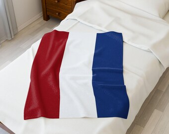 May include: A rectangular blanket with a design of three vertical stripes: red, white, and blue. The blanket is folded and placed on a white bed, with a wooden nightstand and a book visible in the background. The blanket's colors are vibrant.