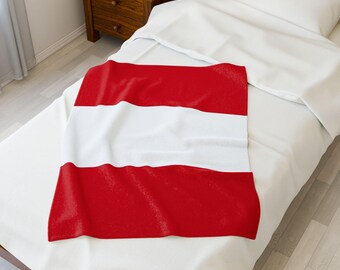 May include: A rectangular red and white blanket, draped over a bed. The blanket features a design with three horizontal stripes: two red stripes at the top and bottom, and a white stripe in the middle. The bed is covered with a white sheet.