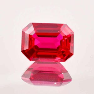 May include: A vibrant red gemstone with a rectangular emerald cut. The gemstone is faceted and reflects light.