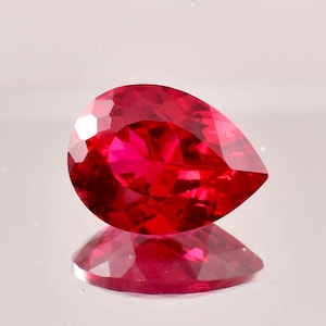 Pode incluir: Uma gema vermelha vibrante em forma de pêra com um corte facetado. A gema reflete a luz, mostrando seu brilho.