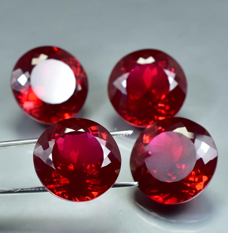 Pode incluir: Quatro pedras preciosas redondas, facetadas, de cor vermelha. As pedras preciosas s&atilde;o engastadas em um material transparente e reflexivo.